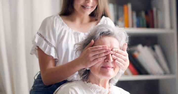 cheerful teenager playing with grandmother guess who game while making surprise in light living room